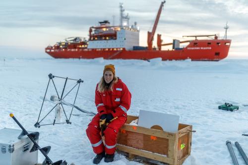Friederike Krüger auf einer Eisscholle im roten, warmen Schneeanzug, im Hintergrund ist das Begleitschiff "Akademik Fedorov" zu sehen.© Quelle: Mario Hoppmann